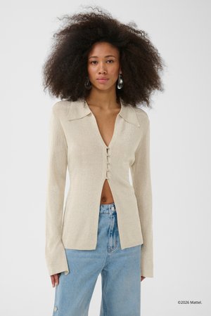 Kobieta o bujnych kręconych włosach, ubrana w jasnobeżowy kardigan zapinany na guziki oraz jasnoniebieskie, wysokowcześnie jeansy, zwrócona twarzą do przodu.