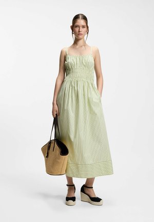 Junge Frau in einem ärmellosen, grün gestreiften Maxikleid, hält eine Strohtasche und trägt schwarze Keil-Espadrilles.