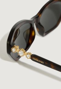 Templi degli occhiali da sole tartarugati con cerniera dorata e tre cristalli Swarovski trasparenti, con il logo "SWAROVSKI" in lettere dorate.