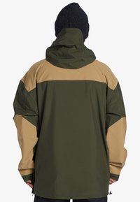 Chaqueta de gran tamaño en verde oscuro y marrón claro, con capucha, panel superior en contraste y textura suave. Incluye bolsillos laterales con cremallera.