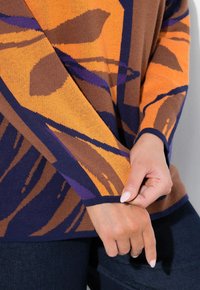 Pull en tricot avec un motif abstrait orange, marron et violet, agrémenté d'un poignet ajusté bleu marine. Gros plan sur la manche et la main.
