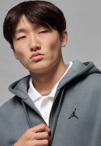 Felpa grigia con zip, caratterizzata da un logo Jumpman nero sul petto sinistro, indossata sopra una maglietta polo bianca, con una texture liscia e un cappuccio.