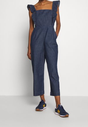 Femme portant une combinaison en denim foncé sans manches avec des bretelles à volants et des baskets bleu marine avec des touches jaunes, debout devant un fond uni.