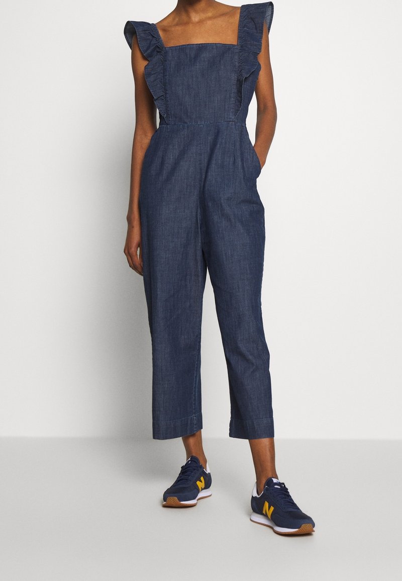 Femme portant une combinaison en denim foncé sans manches avec des bretelles à volants et des baskets bleu marine avec des touches jaunes, debout devant un fond uni.
