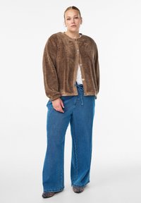 Cardigan ouvert brun et duveteux avec un panneau beige lisse, comportant de grands boutons. Associé à un jean large bleu et des chaussures à imprimé animal.