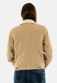 Chaqueta de pana beige con un cuello blanco esponjoso, que presenta un diseño ligeramente entallado y botones a presión ajustables en la cinturilla.