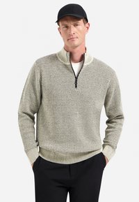 Man in een lichtgrijze, gestructueerde sweater met kwart ritssluiting, zwarte broek en een zwarte baseballpet, met zijn handen in zijn zakken tegen een witte achtergrond.