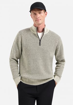 Man in een lichtgrijze, gestructueerde sweater met kwart ritssluiting, zwarte broek en een zwarte baseballpet, met zijn handen in zijn zakken tegen een witte achtergrond.