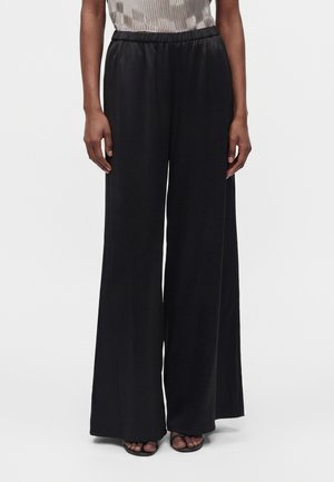 Personne portant un pantalon noir à jambes larges et des sandales noires à bout ouvert, debout devant un fond clair uni.