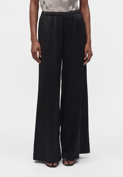 Personne portant un pantalon noir à jambes larges et des sandales noires à bout ouvert, debout devant un fond clair uni.