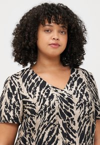 Une jeune femme aux cheveux bouclés et au maquillage naturel regarde la caméra avec confiance, portant une chemise beige et noire à motifs.