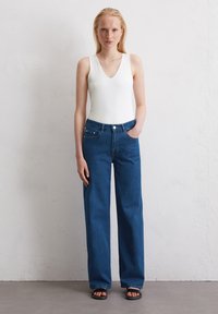 Witte mouwloze bodysuit met een diepe V-hals gecombineerd met hoge taille, wijde donkerblauwe denim jeans. Model staat op een grijze vloer.