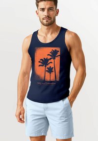 Ein junger Mann trägt ein dunkelblaues Tanktop mit einem orangefarbenen Palmenmotiv und dem Schriftzug "Nimm mich mit ins Paradies", kombiniert mit hellblauen Shorts.