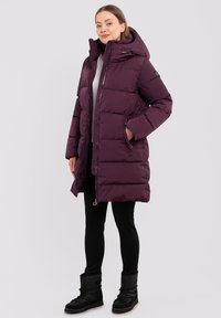 Lila Daunenjacke mit Kapuze, Reißverschluss und Seitentaschen. Weicher Stoff mit einem gesteppten Muster, getragen über schwarzen Leggings und Stiefeln.