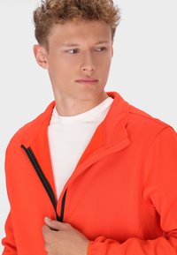 Ryškiai oranžinė zip-up striukė su aukštu apykakle ir juodu užtrauktuku, uždėta ant baltos marškinėlių, pasižyminti lygiu audinio paviršiumi.