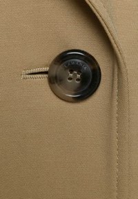 Detailný záber na gombík z korytnačiny prišitý na béžovú textúrovanú tkaninu s našitou dierkou na gombík v blízkosti.