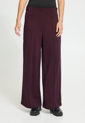 Bordeauxrode broek met wijde pijpen en subtiele verticale strepen, gedragen met zwarte schoenen en een zwart topje, zichtbaar van taille tot beneden.