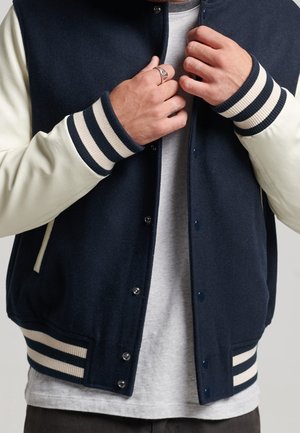 Marineblaue Wolle-Mischung Varsity-Jacke mit cremefarbenen Lederärmeln, gerippten gestreiften Bündchen, Druckknöpfen und Seitentaschen. Graues T-Shirt darunter.
