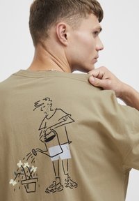Oliven grøn bomulds T-shirt med en sort linjetegning af en person, der vander blomster på ryggen, korte ærmer og rund hals.