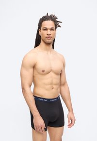 Ing nélküli, izmos férfi hosszú rasztahajjal, fekete Calvin Klein boxeralsót viselve, egyszínű világos háttér előtt áll.