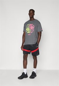 Grå T-shirt med et lyst gul og pink løvegrafik, parret med sorte shorts med røde striber, og sorte sneakers med hvide sokker.
