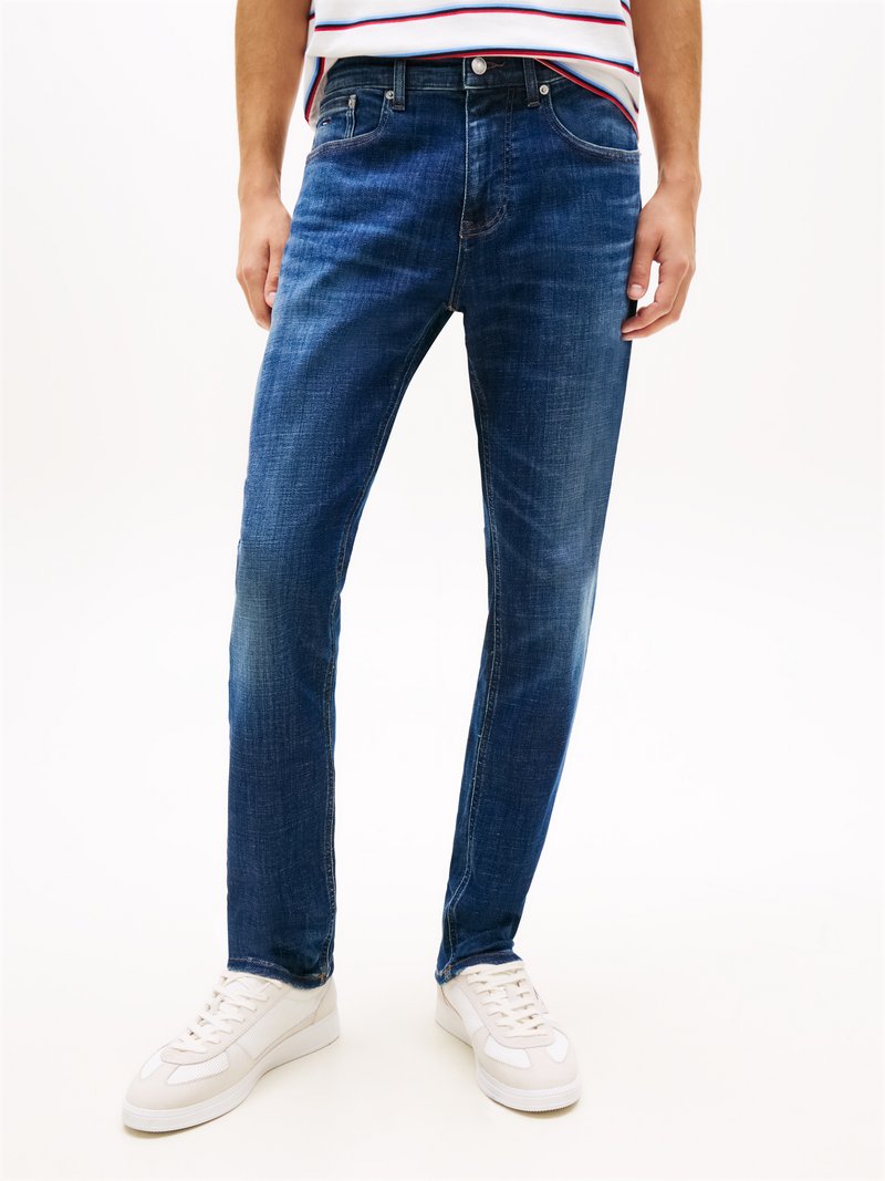Homme portant un jean slim bleu foncé avec une décoloration subtile, des baskets blanches et une chemise rayée, debout devant un fond uni.