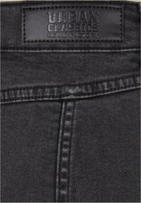 Černá denimová tkanina s hladkou texturou, s koženou etiketou označenou "URBAN CLASSICS DENIM FACTORY" a detailním šitím.
