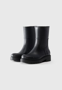 Une paire de bottes en caoutchouc noires mi-mollet avec des semelles épaisses à motif sur un fond clair.