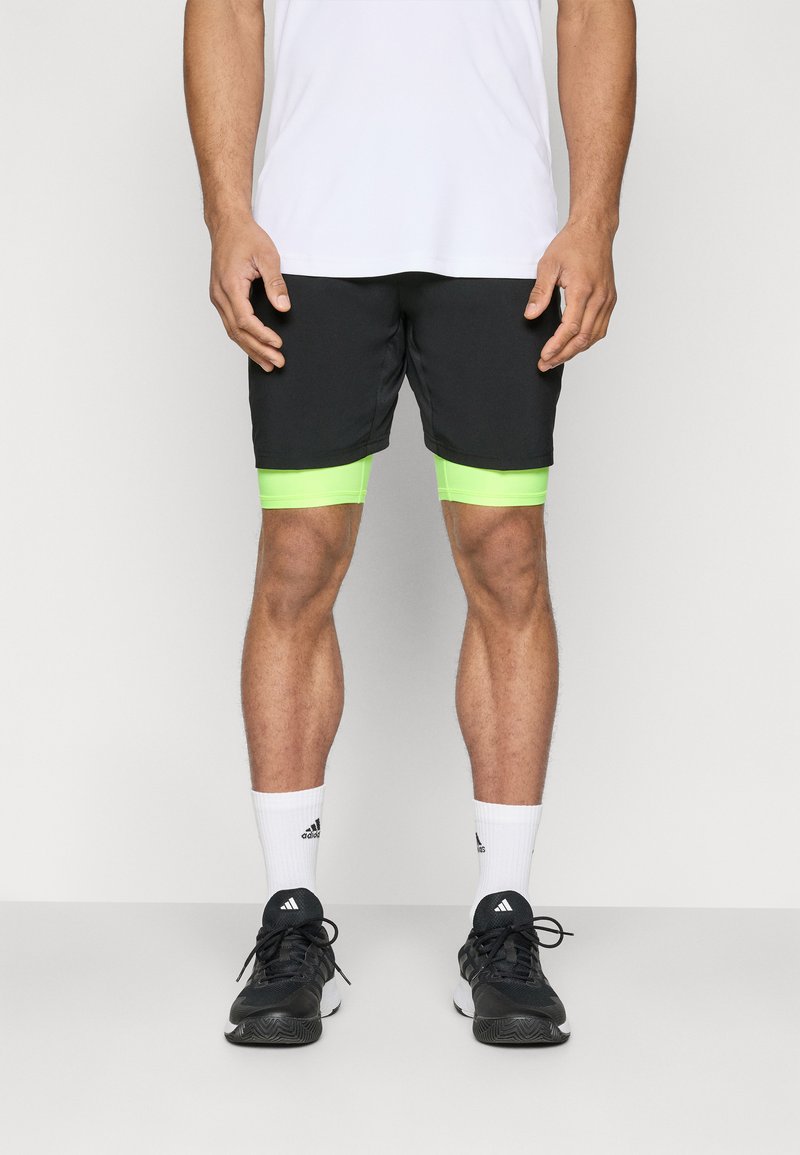 Shorts de sport noirs avec des accents vert fluo, associés à des chaussettes blanches et des baskets noires. Modèle debout dans une pose neutre.