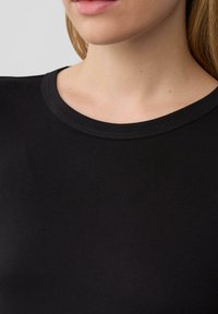 Frau trägt ein schlichtes schwarzes Rundhals-T-Shirt, zeigt Hals, Kinn und einen Teil der schulterlangen blonden Haare.