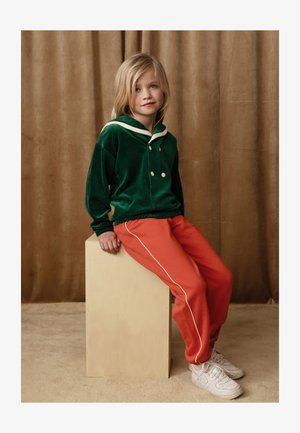 Grønn fløyels genser med hvit krage og knapper, parret med oransje joggebukser med hvite sidestriper, iført hvite sneakers.