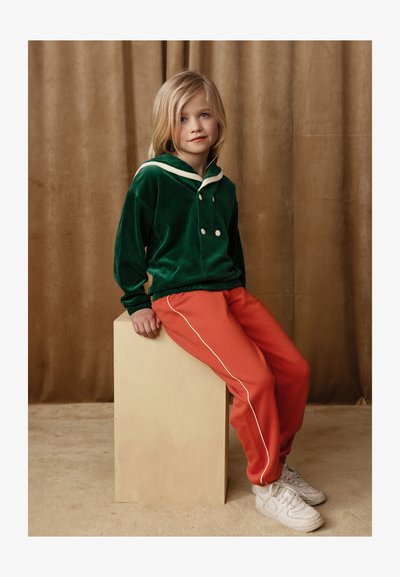 Grön sammetsliknande tröja med vit krage och knappar, i kombination med orangea sweatpants med vita sidostreck, bärs med vita sneakers.