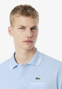 Ljusblå pikétröja i texturerad tyg med vit krage och detaljer. Har en liten broderad krokodillogo på vänster bröst.