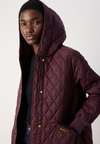 Gestikte bordeauxjas met een capuchon, zichtbare gouden hardware en een zijvlakkenzak met het "Barbour" logo. Gewoven stof en moderne snit.