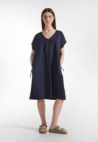 Marineblaues Kleid mit V-Ausschnitt und kurzen Ärmeln, ausgestattet mit elastischem Bund und Seitentaschen; strukturierter Stoff. Kombiniert mit braunen Sandalen.