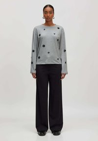 Vrouw staat op met een grijze lange mouwen top met zwarte bloemenpatches, zwarte wijde broek, zwarte schoenen en zilveren creolen.