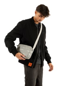 Borsa a tracolla grigia con superficie strutturata, caratterizzata da stampa del logo e tracolla resistente; portata da una persona con giacca bomber nera.