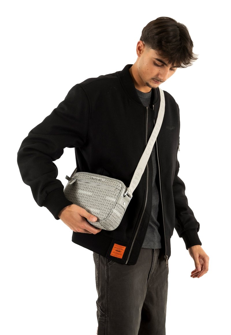 Borsa a tracolla grigia con superficie strutturata, caratterizzata da stampa del logo e tracolla resistente; portata da una persona con giacca bomber nera.