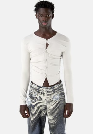 Uomo con indosso un top bianco aderente, arricciato, con bottoni e maniche lunghe e jeans consumati marmorizzati nere e bianche, in piedi davanti a uno sfondo semplice.