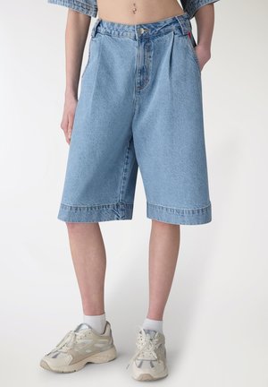 Persona che indossa pantaloncini Bermuda in denim azzurro chiaro, calzini bianchi e scarpe da ginnastica beige, in piedi contro uno sfondo bianco neutro.