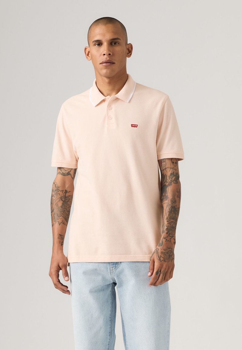 Jeune homme avec des tatouages portant un polo Levi's rose clair et un jean bleu clair sur un fond uni.