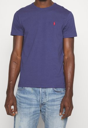 Mann trägt ein schlichtes blaues Kurzarm-T-Shirt mit einem kleinen roten gestickten Logo auf der linken Brust und hellblaue ausgewaschene Jeans.