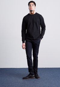 Chemise noire à manches longues, design ajusté, associée à un pantalon sombre et des baskets noires. Texture lisse, sans motifs ni logos visibles.