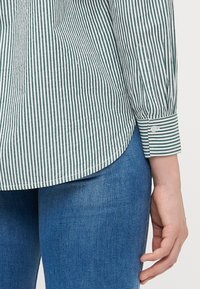 Grønn og hvit stripete skjorte med lange ermer, knappede mansjetter og buet nederkant, kombinert med blå jeans. Stoffet ser glatt ut.
