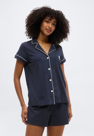 Ženska s kodrastimi lasmi v mornarsko modri kratkih rokavih srajci za pižamo z gumbi in ujemajočih kratkih hlačah z belimi pikami in obrobami.