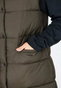 Olivgrüner, gepolsterter Weste mit gestepptem Muster, ausgestattet mit schwarzen Knöpfen, einer Seitentasche und einem blauen Langarmshirt darunter.