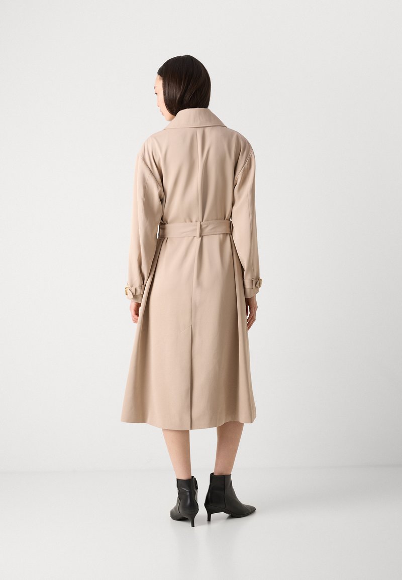 Beiger Trenchcoat mit tailliertem Kleidungsstil, langen Ärmeln und einem Schlitz im Rücken. Glattes Material und maßgeschneiderte Passform. Schwarze Knöchelschürzen runden den Look ab.