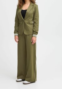 Costume vert olive comprenant un blazer ajusté à bouton unique et un pantalon à jambes larges avec des bandes blanches sur les côtés, assorti de chaussures claires.