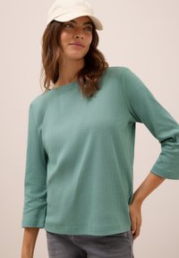 Femme portant un haut à manches longues côtelé vert clair, un jean gris et une casquette de baseball blanche, posant avec une main sur la hanche.