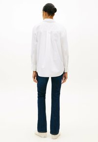 Mujer con un moño oscuro, lleva una camisa blanca holgada de botones, vaqueros rectos azul oscuro y zapatos blancos, de pie frente a un fondo blanco.